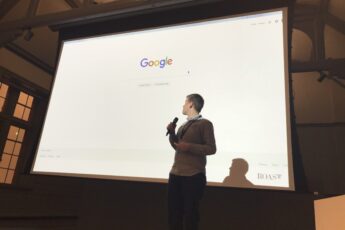 Man holding a microphone stands in front of a large projection screen displaying the Google homepage during a presentation or conference talk. The Google logo and search bar are visible on the screen, with the word ROAST shown in the bottom corner, highlighting a digital marketing or SEO focused event setting.