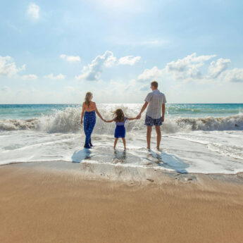 A family of three stands hand in hand at the edge of the ocean as waves roll onto a sandy beach under a bright blue sky. The parents and child face the water together, capturing a joyful seaside vacation moment and the feeling of spending quality time together by the sea.