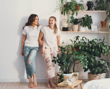 Two women wearing casual white T shirts and ripped jeans stand barefoot against a white wall, smiling and laughing together in a bright indoor space. Beside them, wooden shelves are filled with lush green houseplants in pots, creating a cozy and modern home setting that highlights indoor plant decor and relaxed lifestyle vibes.