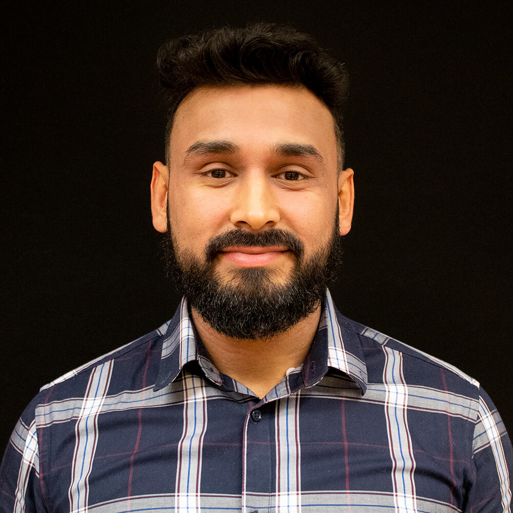 Close up headshot of a man with short dark hair and a full beard wearing a navy and white plaid button down shirt against a black background. He faces the camera with a calm slight smile, creating a professional and approachable portrait suitable for a team bio or company about page.