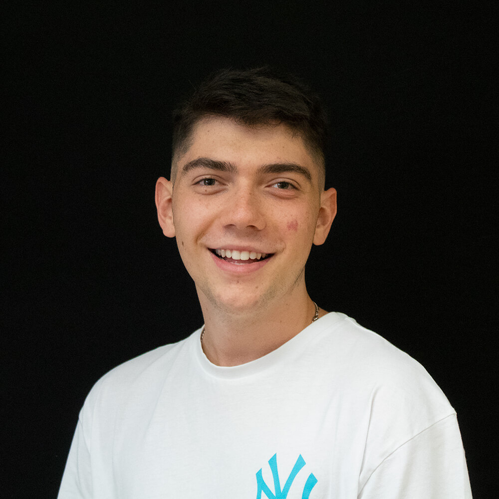 Close up headshot of a young man with short dark hair wearing a white T shirt against a black background. He smiles directly at the camera, creating a friendly and approachable portrait suitable for a team member profile or about page.