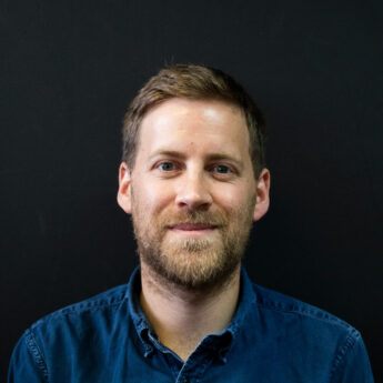 Close up headshot of a man with short light brown hair and a neatly trimmed beard wearing a dark blue button down shirt against a black background. He looks directly at the camera with a subtle smile, creating a professional and approachable portrait suitable for a team bio or company about page.