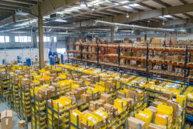 Large warehouse interior with tall shelving units, yellow bins, and stacked cardboard boxes organized for shipping and storage.