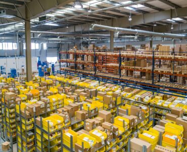 Large warehouse interior with tall shelving units, yellow bins, and stacked cardboard boxes organized for shipping and storage.