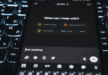 A person using a smartphone to interact with the ChatGPT app, the screen showing options like “Create images,” “Code,” and “Analyse data,” with a softly lit monitor blurred behind it.