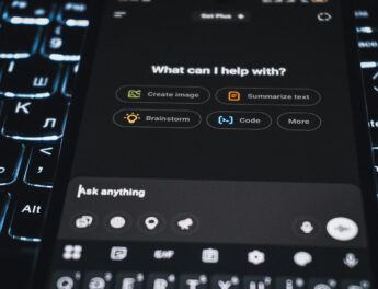 A person using a smartphone to interact with the ChatGPT app, the screen showing options like “Create images,” “Code,” and “Analyse data,” with a softly lit monitor blurred behind it.