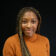 Smiling woman with long brown hair wearing an orange turtleneck jumper, posed against a black background.