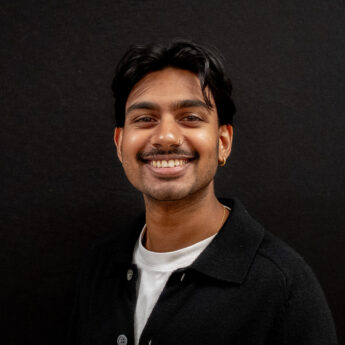 Close up headshot of a man with short dark hair and a trimmed mustache wearing a black jacket over a white shirt against a black background. He smiles broadly at the camera, creating a polished and approachable portrait suitable for a team member profile or about page.