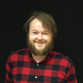 Close up headshot of a man with medium length brown hair and a full beard wearing a red and black plaid button down shirt against a black background. He smiles warmly at the camera, creating a friendly and approachable portrait suitable for a team bio or about page.