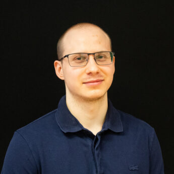 Close up headshot of a man with very short light hair wearing glasses and a navy polo shirt against a black background. He looks directly at the camera with a neutral expression, creating a clean and professional portrait suitable for a team member profile or company about page.