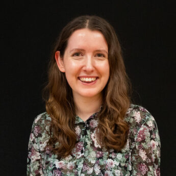 Smiling woman with long brown hair wearing a floral blouse, posed against a black background.