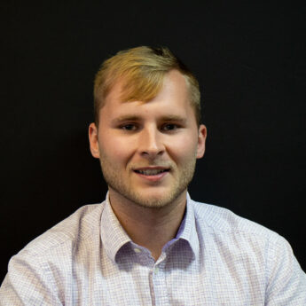 Close up headshot of a man with short blond hair and light stubble wearing a light patterned button down shirt against a black background. He looks directly at the camera with a slight smile, creating a professional and approachable portrait suitable for a team bio or company about page.
