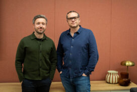 Two adult men standing side by side in a modern interior against a textured terracotta wall, one wearing a dark green button-up jacket and the other in a navy blue shirt with glasses, both casually posed with hands in pockets; minimalist wooden console table beside them holding a striped ceramic vase and a brass table lamp, conveying a professional portrait, contemporary office setting, and business team aesthetic.