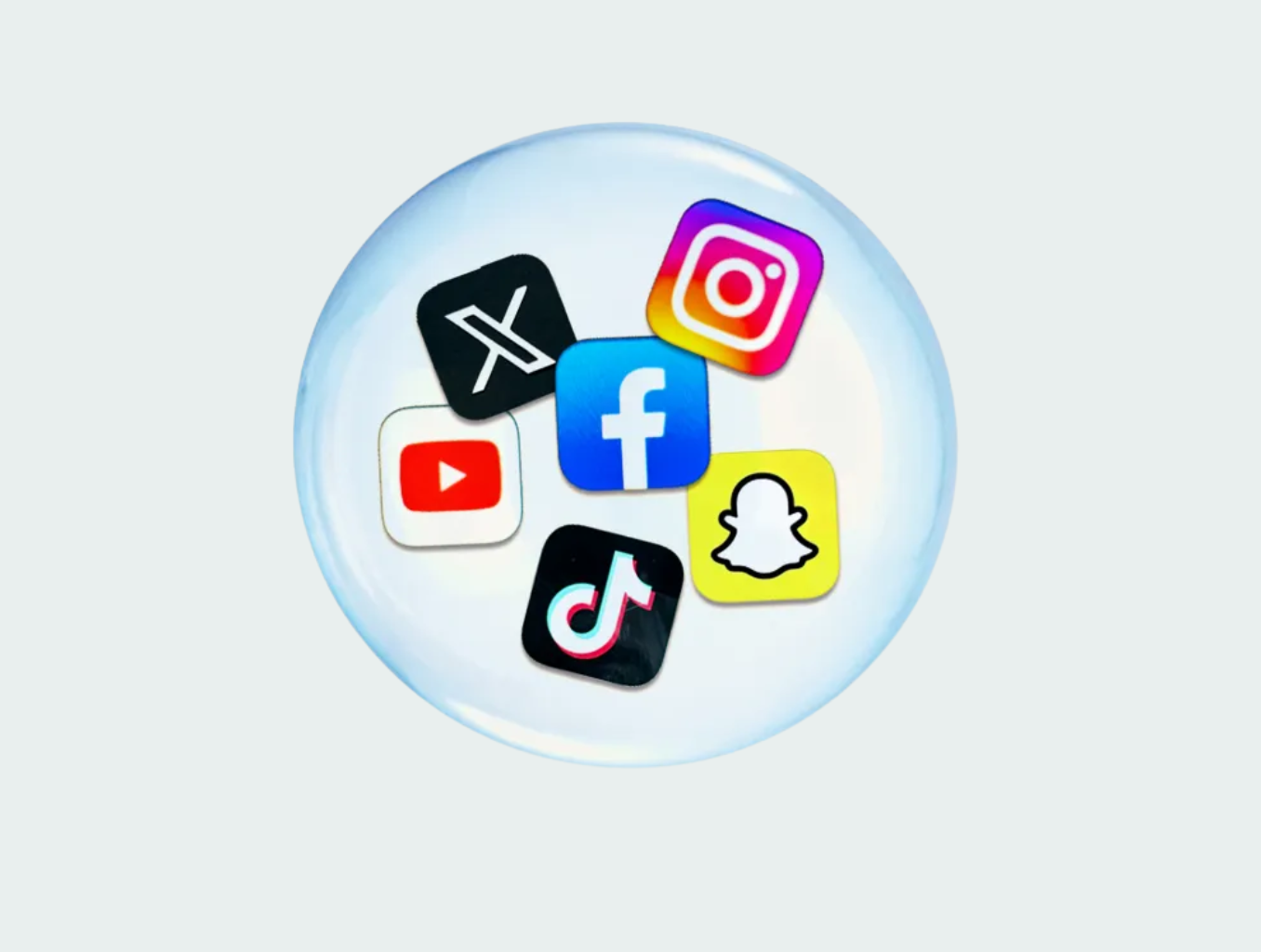 Six colorful social media platform icons arranged in a circular pattern within a translucent glass sphere: X (formerly Twitter) with a white X on black background in the top left, Instagram with its gradient logo in the top right, Facebook with its blue logo in the center, YouTube with a red play button on white background on the left, TikTok with a pink and white musical note on black background at the bottom, and Snapchat with a yellow ghost icon on the bottom right. The sphere sits on a light gray background with a subtle shadow effect.