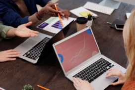 Overhead view of a team meeting around a wooden table, with a laptop in the foreground displaying a line chart and donut chart in red and blue tones, a person pointing at a printed bar chart with an orange pencil, another laptop, a coffee cup, notepad, and small succulent plants nearby.